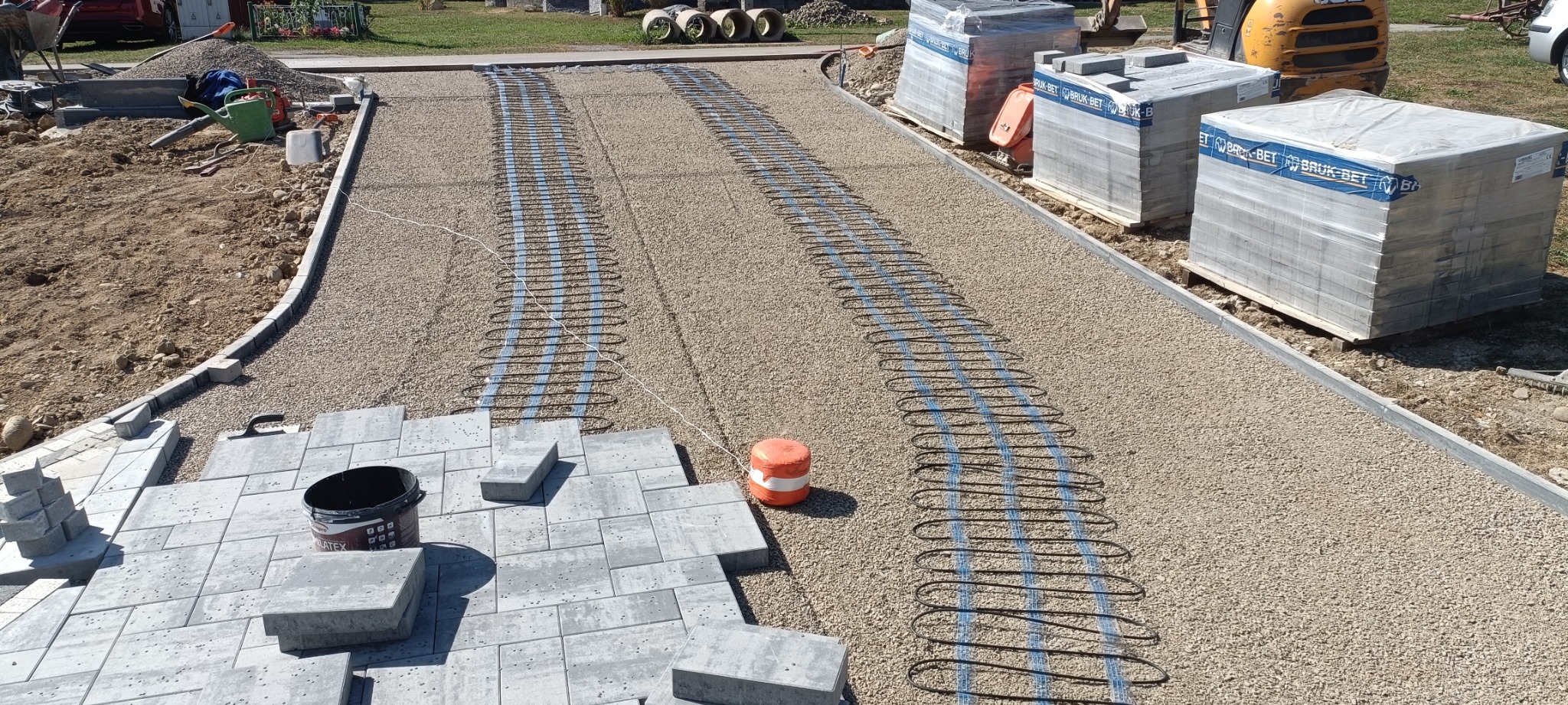 Niedokończony podjazd z szarej kostki brukowej z widocznym systemem ogrzewania podpowierzchniowego na podsypce z kruszywa, palety z kostką Bruk-Bet w tle, widok z góry.