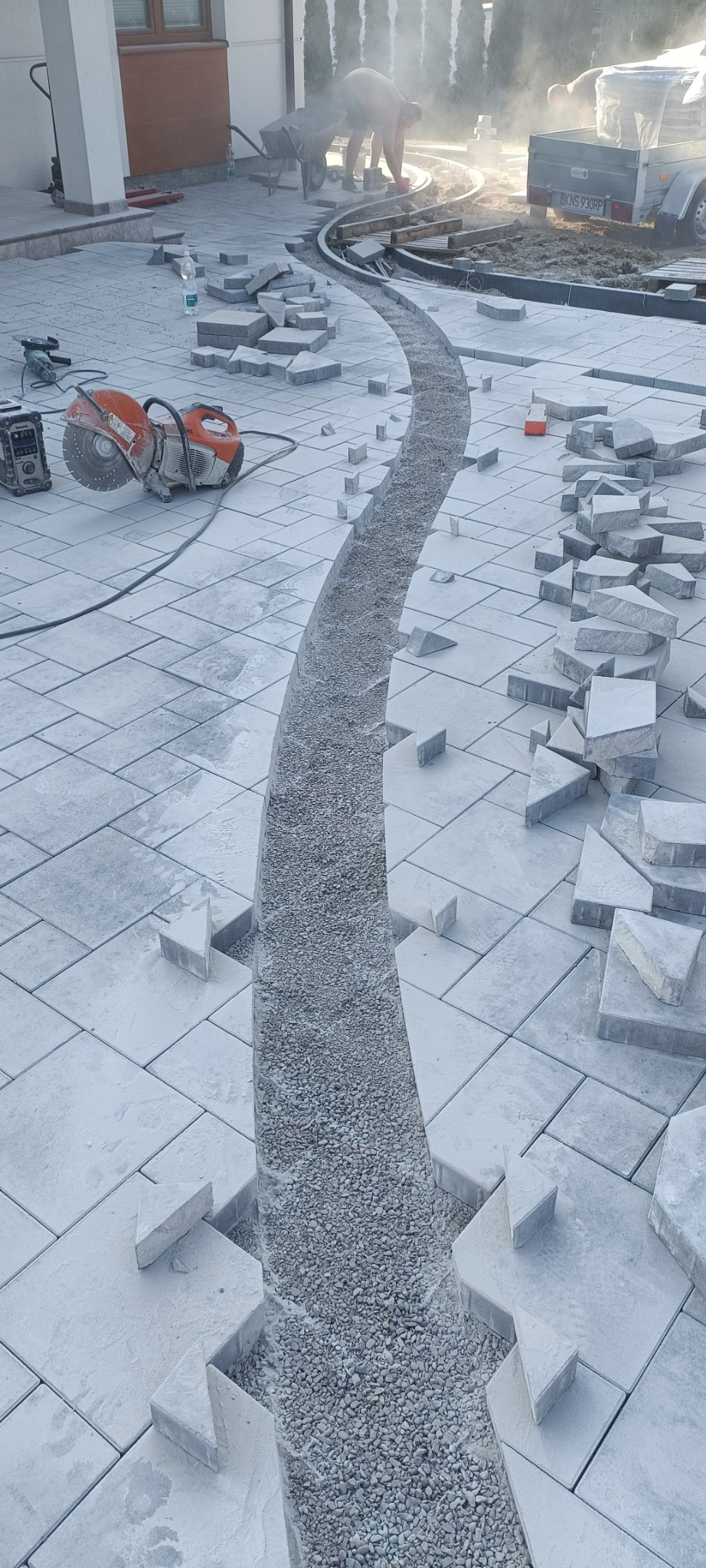Niedokończona ścieżka z szarego żwiru wijąca się przez plac z szarej kostki brukowej, z widocznymi narzędziami do cięcia i robotnikami w tle.