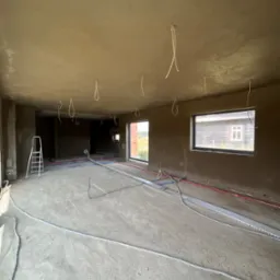 Surowe wnętrze w trakcie remontu: ściany i sufit pokryte świeżym tynkiem, widoczne kable zwisające z sufitu, rury na podłodze, drabina oparta o ścianę, okna z widokiem na zewnątrz.