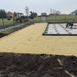 PEDRO-POLAKO Lucas VLASÁK - Przygotowanie terenu pod fundamenty: wyrównany piasek, zbrojenie, pracownik przy pracy. W tle koparka i zabudowa jednorodzinna.