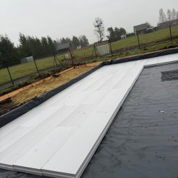 PEDRO-POLAKO Lucas VLASÁK - Ułożone białe płyty izolacyjne na czarnej folii, przygotowane pod fundament. Widoczne ogrodzenie i zieleń w tle, pochmurne niebo.