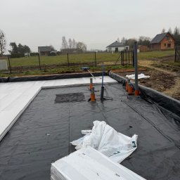 PEDRO-POLAKO Lucas VLASÁK - Przygotowanie podłoża pod fundamenty: czarna folia, izolacja termiczna XPS, rury kanalizacyjne. W tle zabudowa jednorodzinna i ogrodzenie.