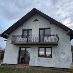 PEDRO-POLAKO Lucas VLASÁK - Dwukondygnacyjny dom z balkonem z kutą balustradą, w trakcie ocieplania elewacji. Widoczne okna z brązowymi ramami i szara, surowa elewacja. Pochmurne niebo w tle.