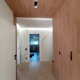 Wykończony korytarz z drewnianą okładziną sufitową i ścienną, białymi drzwiami i podłogą, widok na dalsze pomieszczenie z marmurowymi ścianami i oknem.