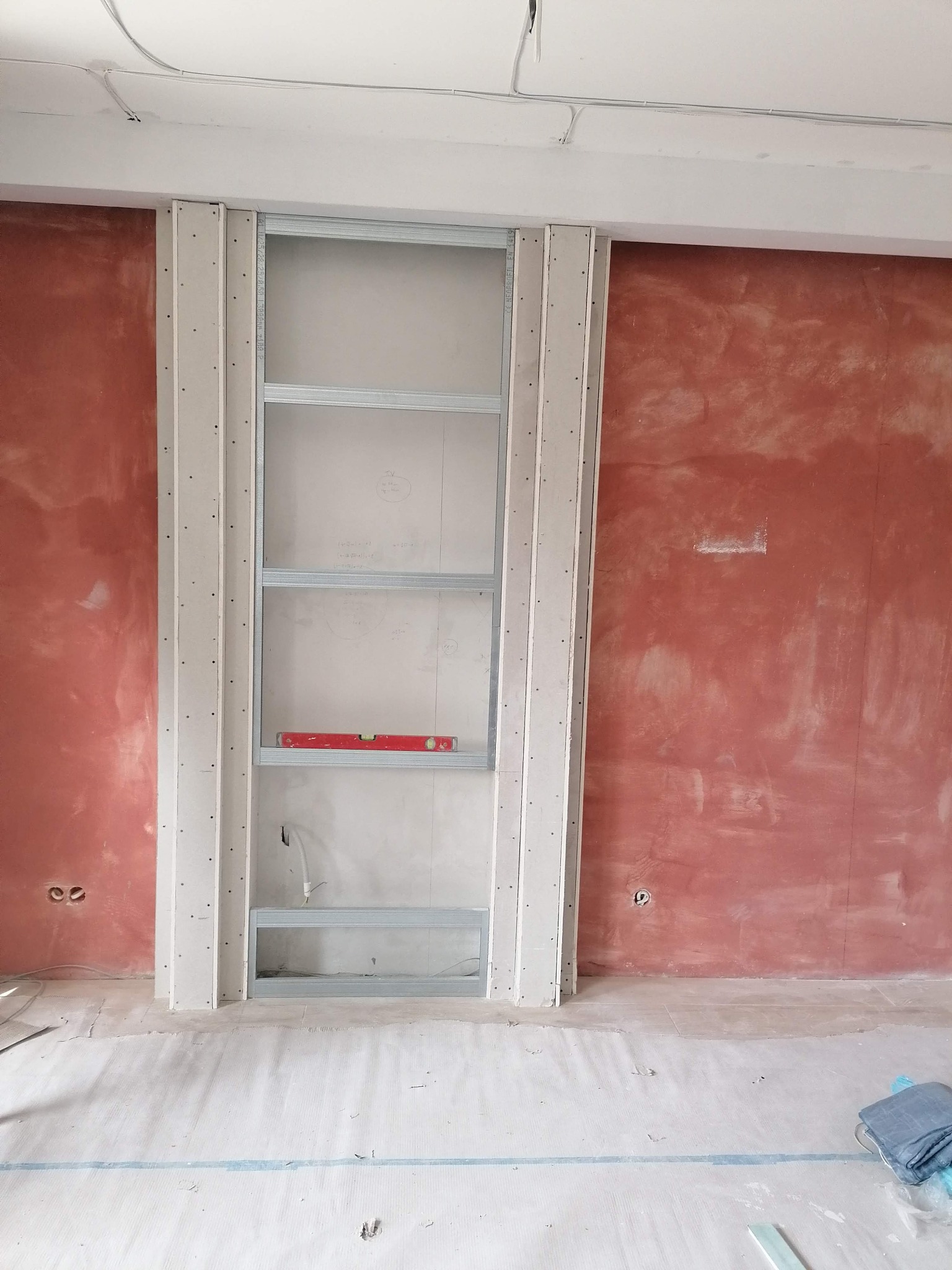 Stelaż z profili metalowych i płyt kartonowo-gipsowych, tworzący wnękę w ścianie z surowym wykończeniem, widoczna poziomica na jednym z poziomów stelażu.