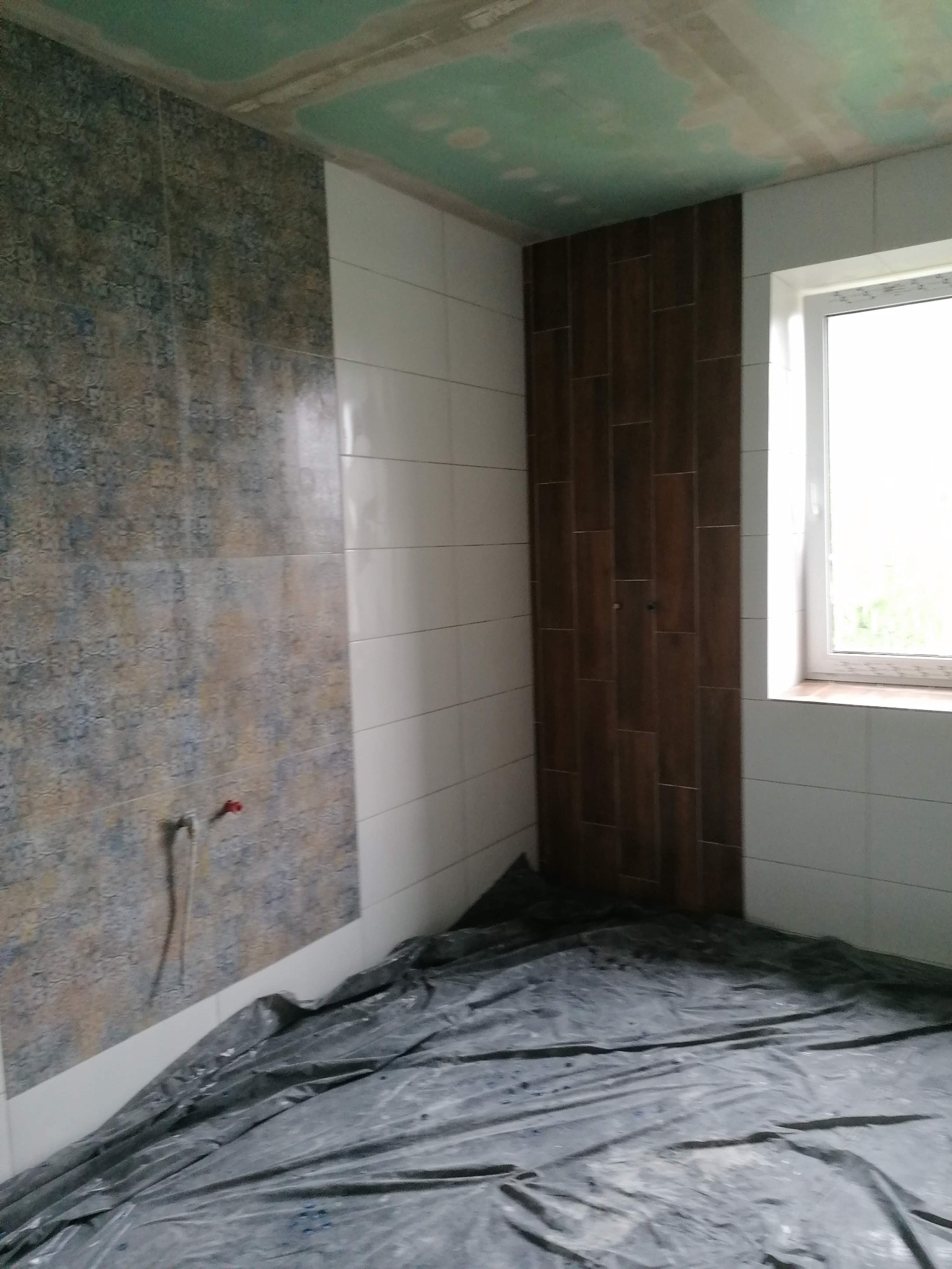 Wnętrze łazienki w trakcie remontu: ściany częściowo wyłożone płytkami w różnych wzorach (imitacja drewna, wzorzyste i białe), podłoga zabezpieczona folią ochronną, widoczne wystające rury...