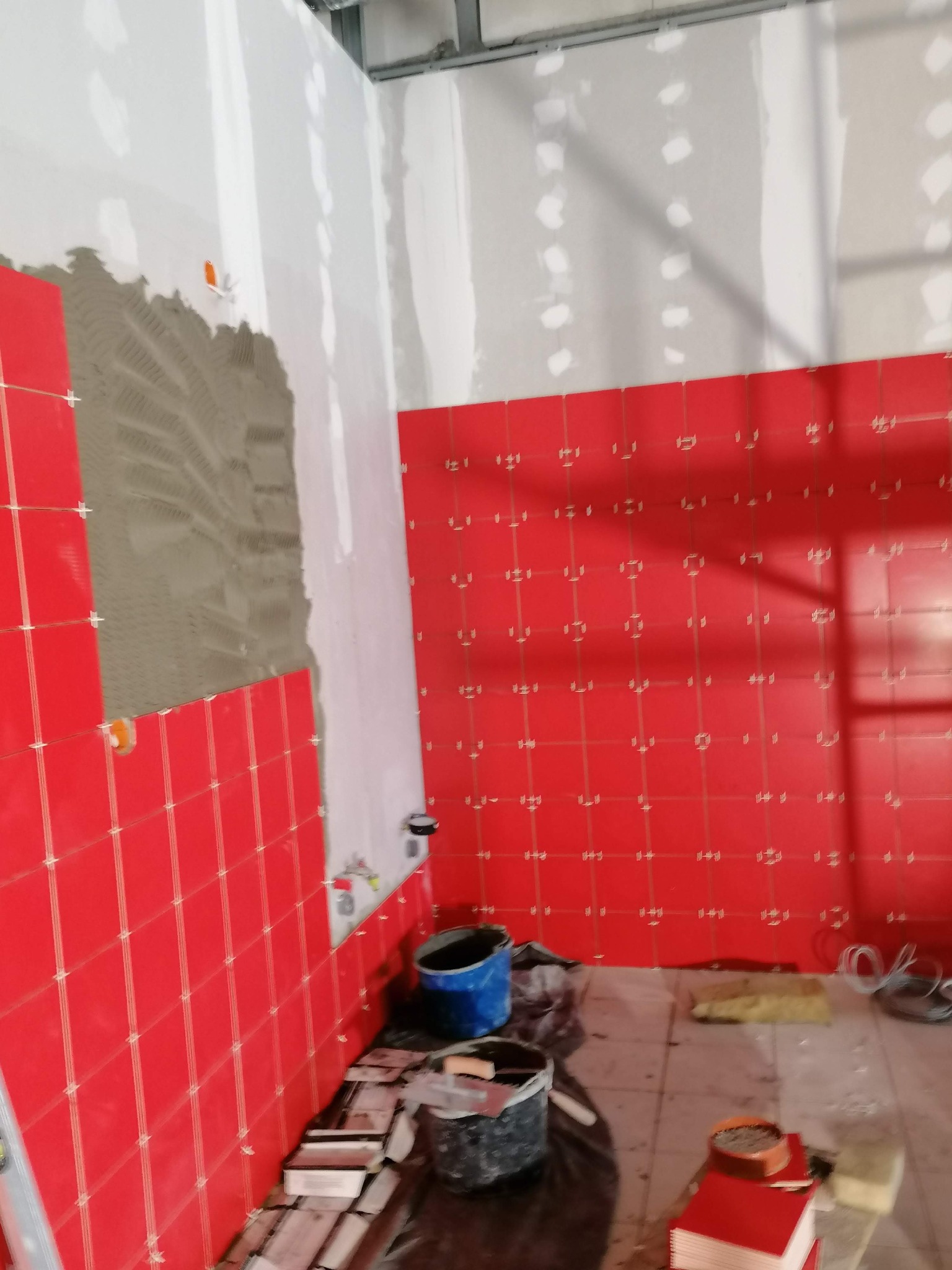 Proces układania czerwonych płytek w łazience, widoczna ściana z nałożonym klejem i częściowo ułożonymi płytkami, a także ułożona ściana z białymi krzyżykami dystansowymi, niebieskie wiadro...
