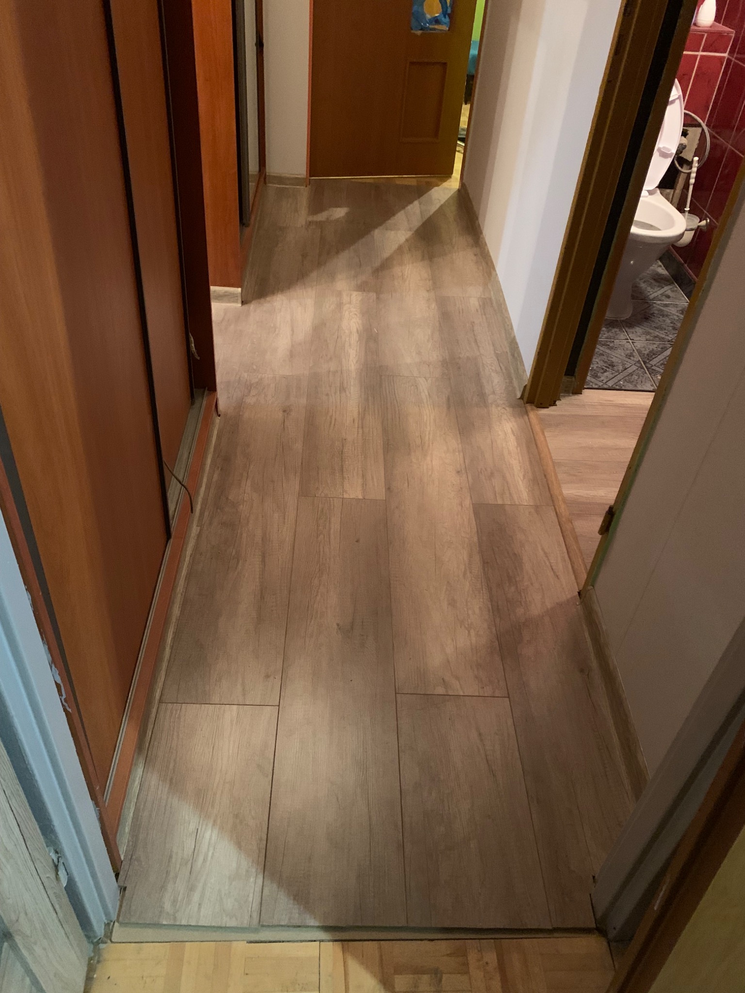 Korytarz z nowo ułożonymi panelami podłogowymi o wzorze drewna, widok z perspektywy drzwi do łazienki, z widoczną toaletą w tle.
