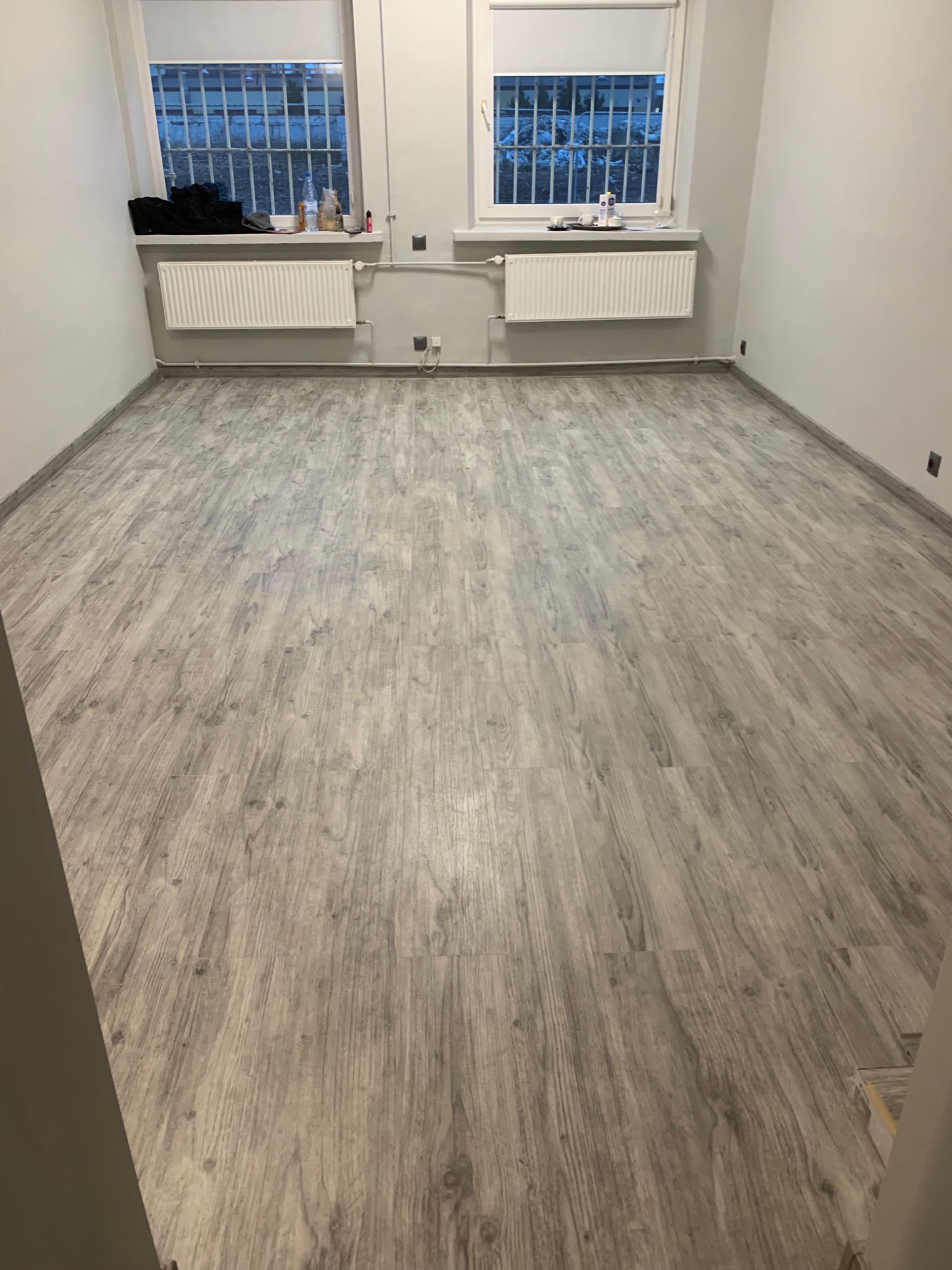 Jasne, szare panele podłogowe z wzorem drewna w pustym pokoju z dwoma oknami z kratami. Na parapetach przedmioty osobiste i grzejniki.