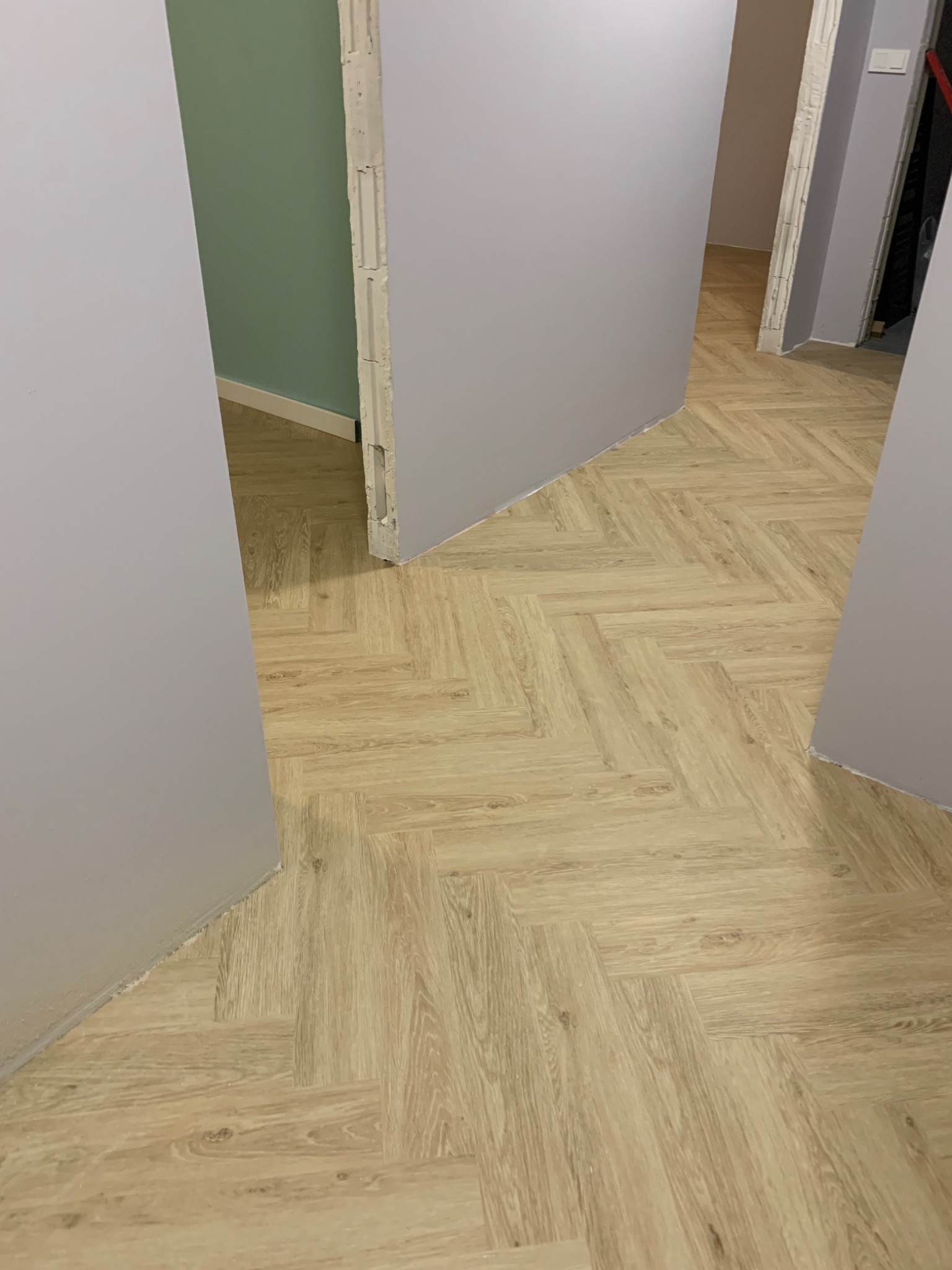 Jasne panele ułożone we wzór jodełki w korytarzu z szarymi ścianami i widocznymi ościeżnicami.