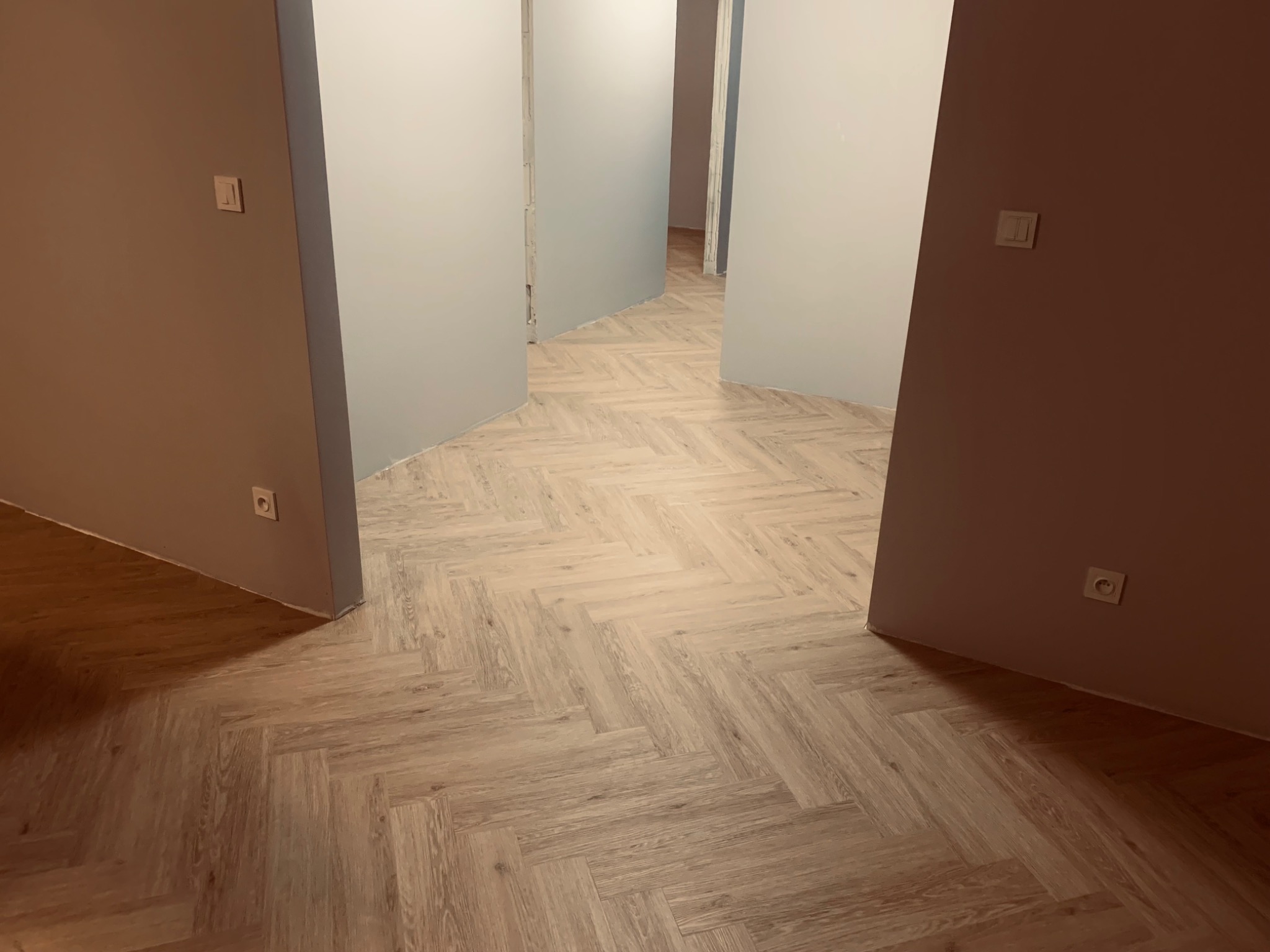 Jasny parkiet ułożony we wzór jodełki francuskiej w pustym, pomalowanym na szaro pomieszczeniu z widocznymi gniazdkami i włącznikami światła.