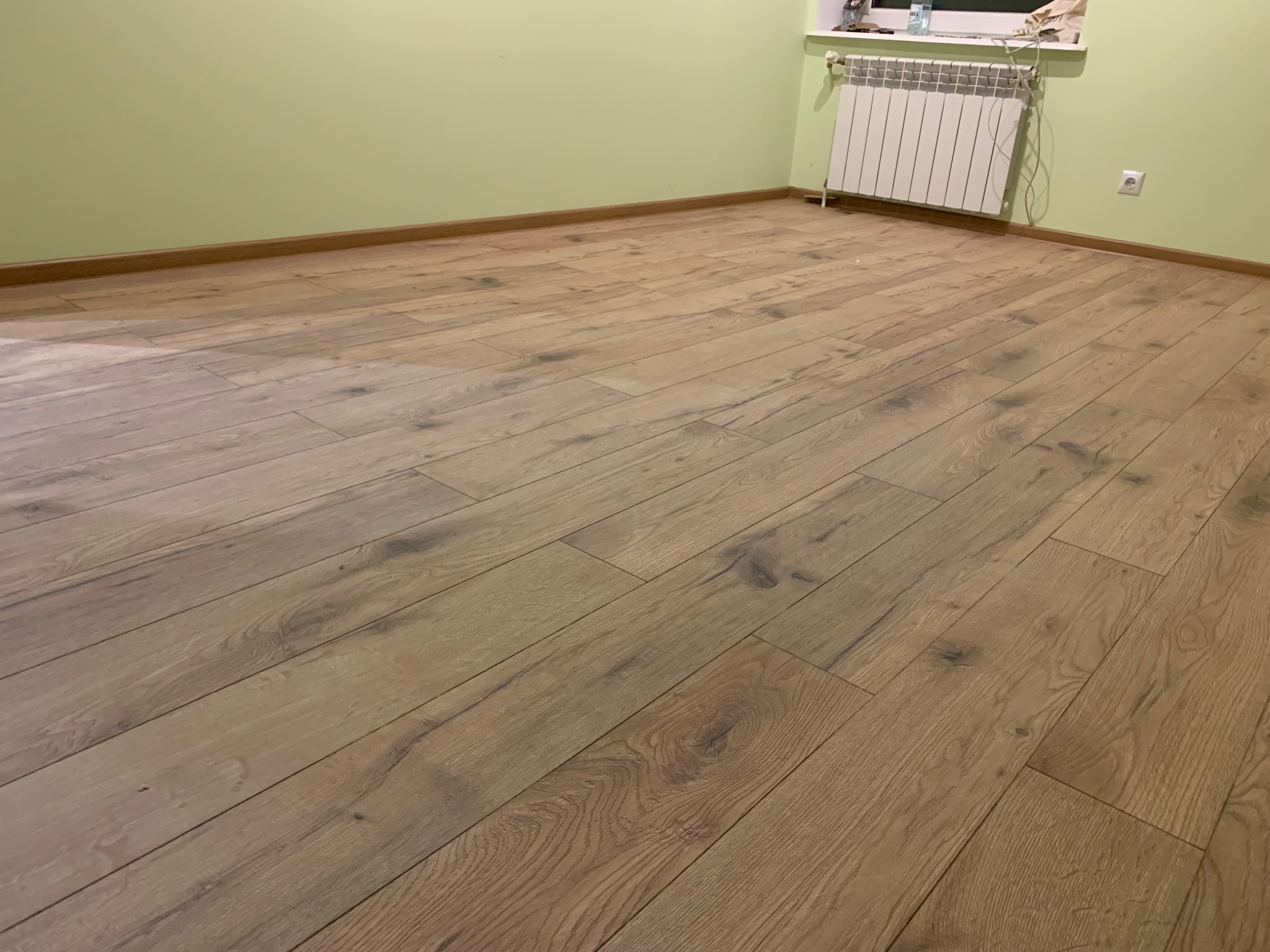 Ukończona podłoga z paneli w odcieniach brązu i beżu, ułożona w jasnym pokoju z zielonymi ścianami i białym grzejnikiem pod oknem.