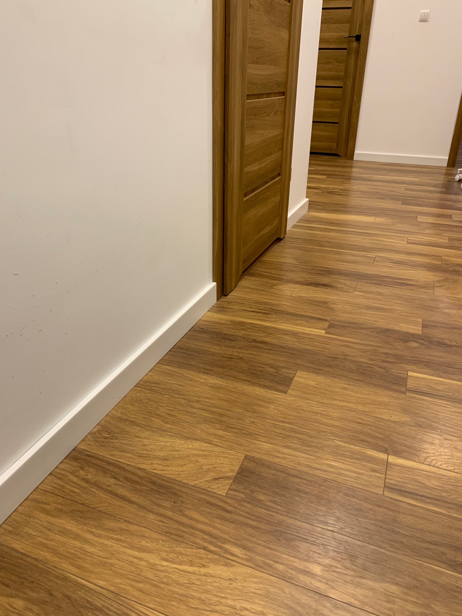 Ułożona podłoga z paneli imitujących drewno, widoczne białe listwy przypodłogowe i fragment drewnianej futryny drzwiowej.