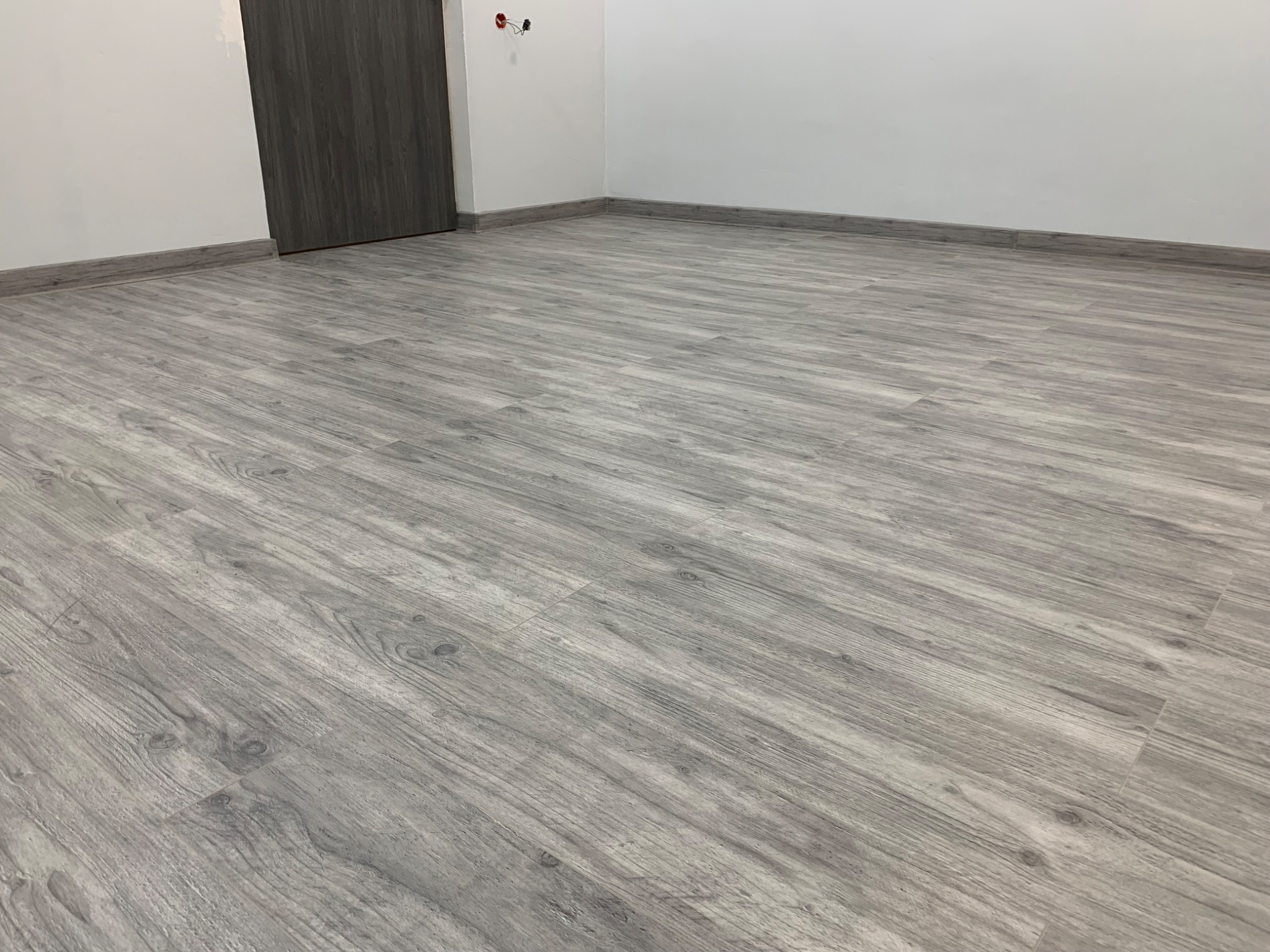 Szare panele podłogowe imitujące drewno, ułożone w pustym pokoju z białą ścianą i szarą listwą przypodłogową, widoczne gniazdka elektryczne nad drzwiami.