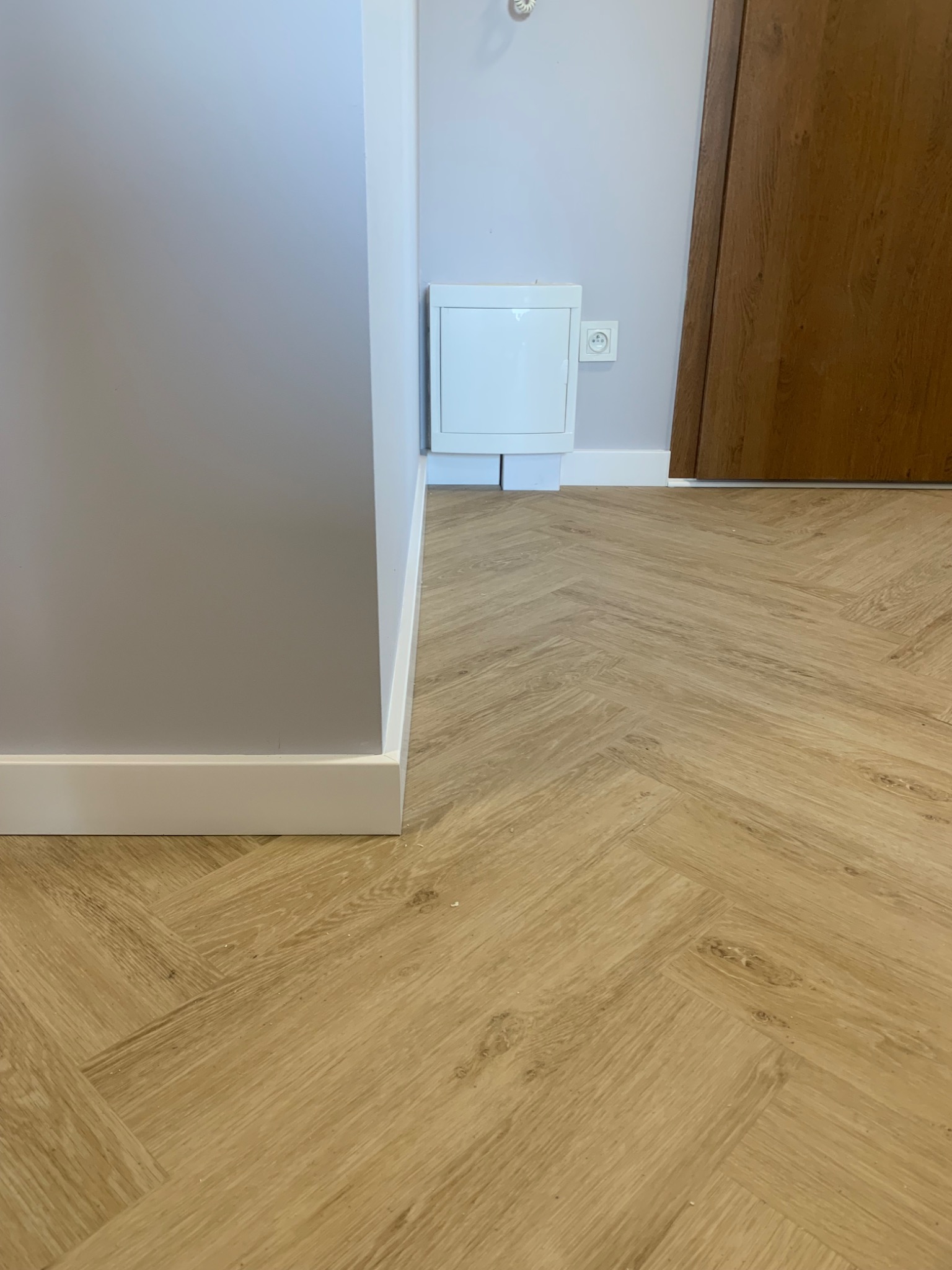 Jasny parkiet ułożony we wzór jodełki francuskiej, widoczny fragment szarej ściany z białą listwą przypodłogową oraz biała szafka z gniazdkiem elektrycznym obok brązowych drzwi.