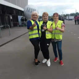 Trzy kobiety w żółtych kamizelkach odblaskowych stoją przed stadionem, uśmiechają się i pokazują kciuki w górę, widoczny balon z napisem GROW.