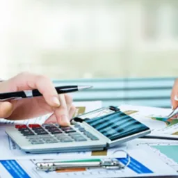 Dwie osoby pracują nad dokumentami finansowymi przy biurku, jedna używa kalkulatora, a druga pisze długopisem, analizując wykresy.