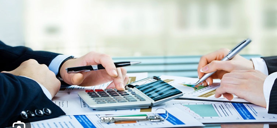 Dwie osoby pracują nad dokumentami finansowymi przy biurku, jedna używa kalkulatora, a druga pisze długopisem, analizując wykresy.