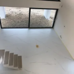 Jasna, marmurkowa podłoga w nowoczesnym wnętrzu z widokiem na piasek przez duże okno, schody w lewym dolnym rogu, żółta taśma malarska przy ścianie.