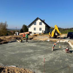 J-kop, prace ziemne Jakub Gręda - Wylany beton pod fundamenty domu jednorodzinnego, widoczna żółta koparko-ładowarka JCB, ubijarka, pracownicy, wykopane fundamenty, niebieskie niebo i krajobraz w tle.