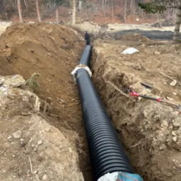 Długi odcinek czarnej, karbowanej rury ułożonej w głębokim wykopie w ziemi, zabezpieczony workami; w tle widoczna postać w ciemnym ubraniu i drzewa.