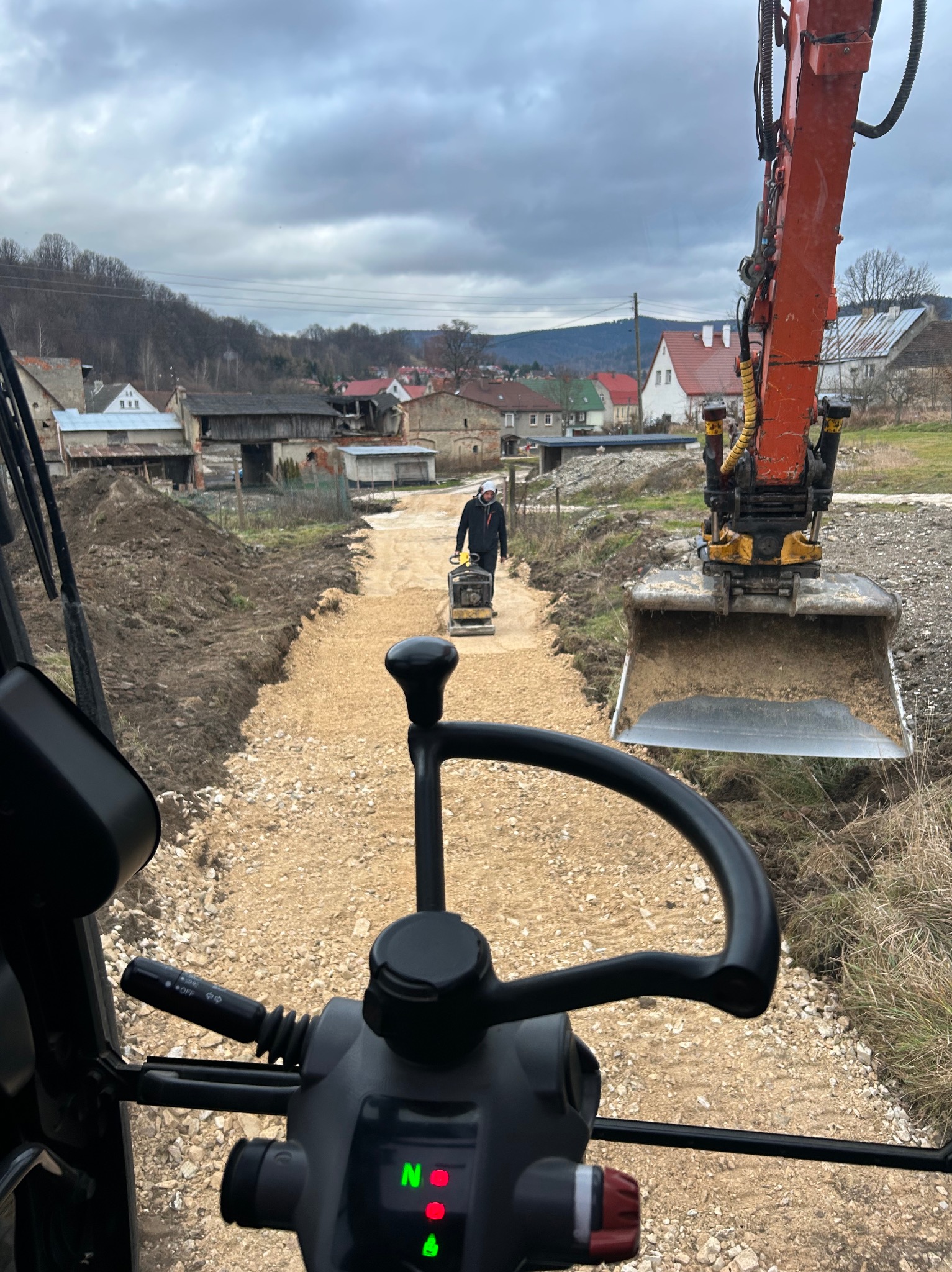 Widok z kabiny koparki na wyrównywany teren pod budowę drogi; w oddali pracownik obsługuje zagęszczarkę, w tle zabudowania wiejskie i zalesione wzgórza.