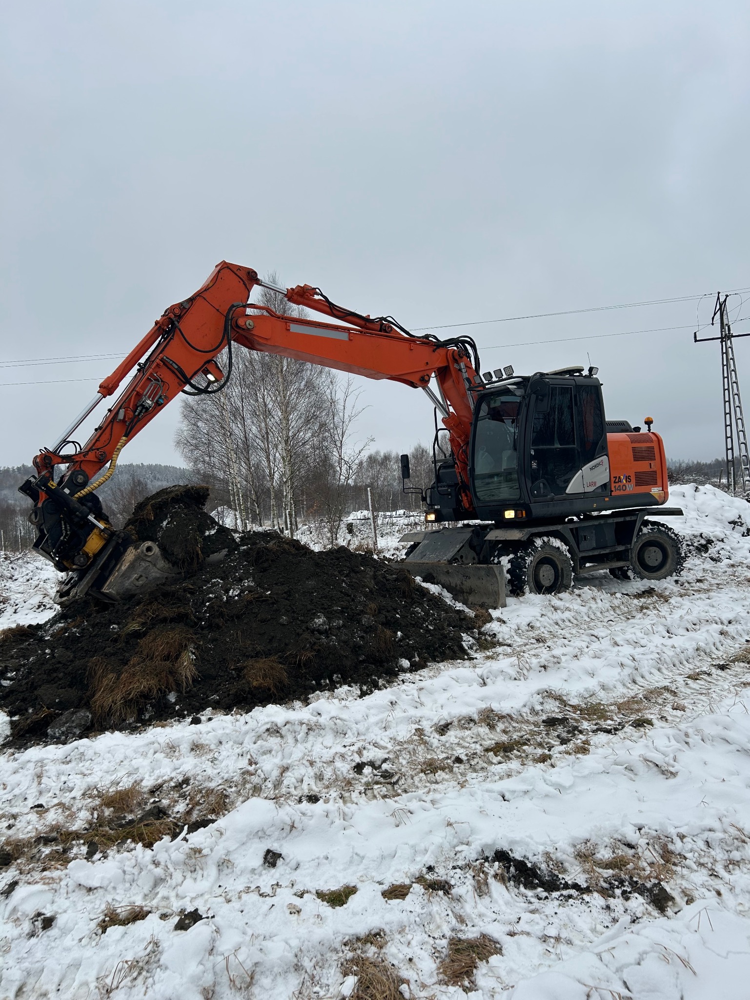 Pomarańczowa koparko-ładowarka Zaxis 140W nabiera łyżką ziemię na zaśnieżonym terenie w pochmurny dzień.