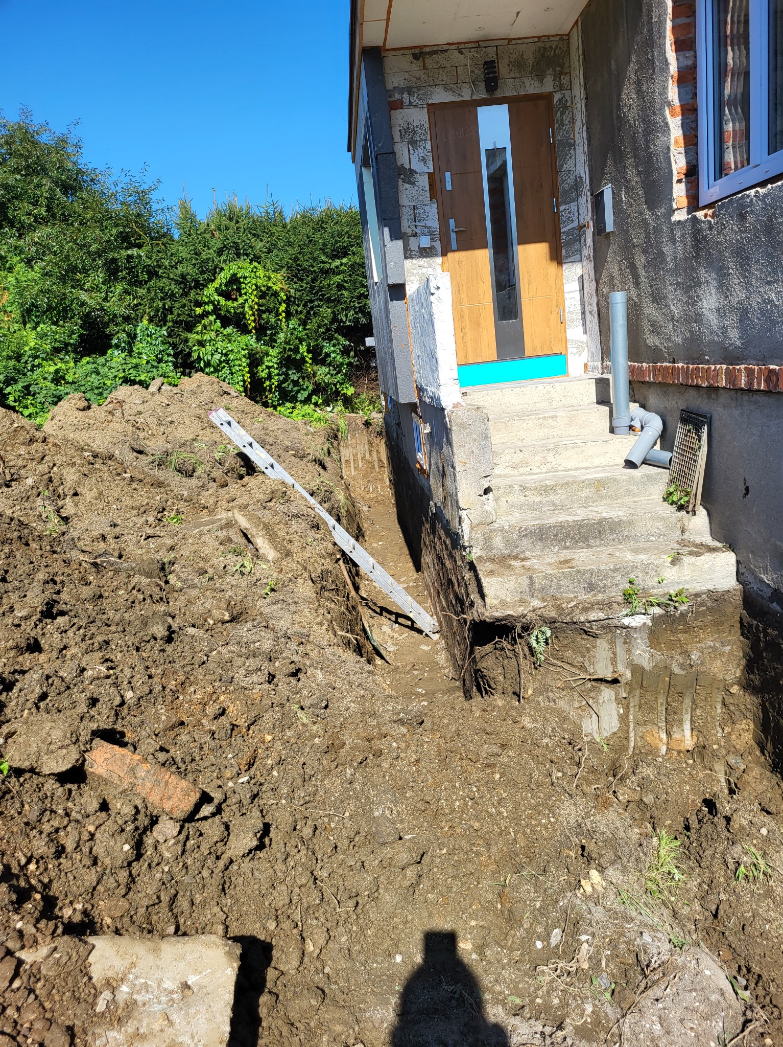 Wykop ziemny odsłaniający fundamenty schodów wejściowych do budynku, z widoczną drabiną opartą o pryzmę ziemi i fragmentem instalacji kanalizacyjnej.