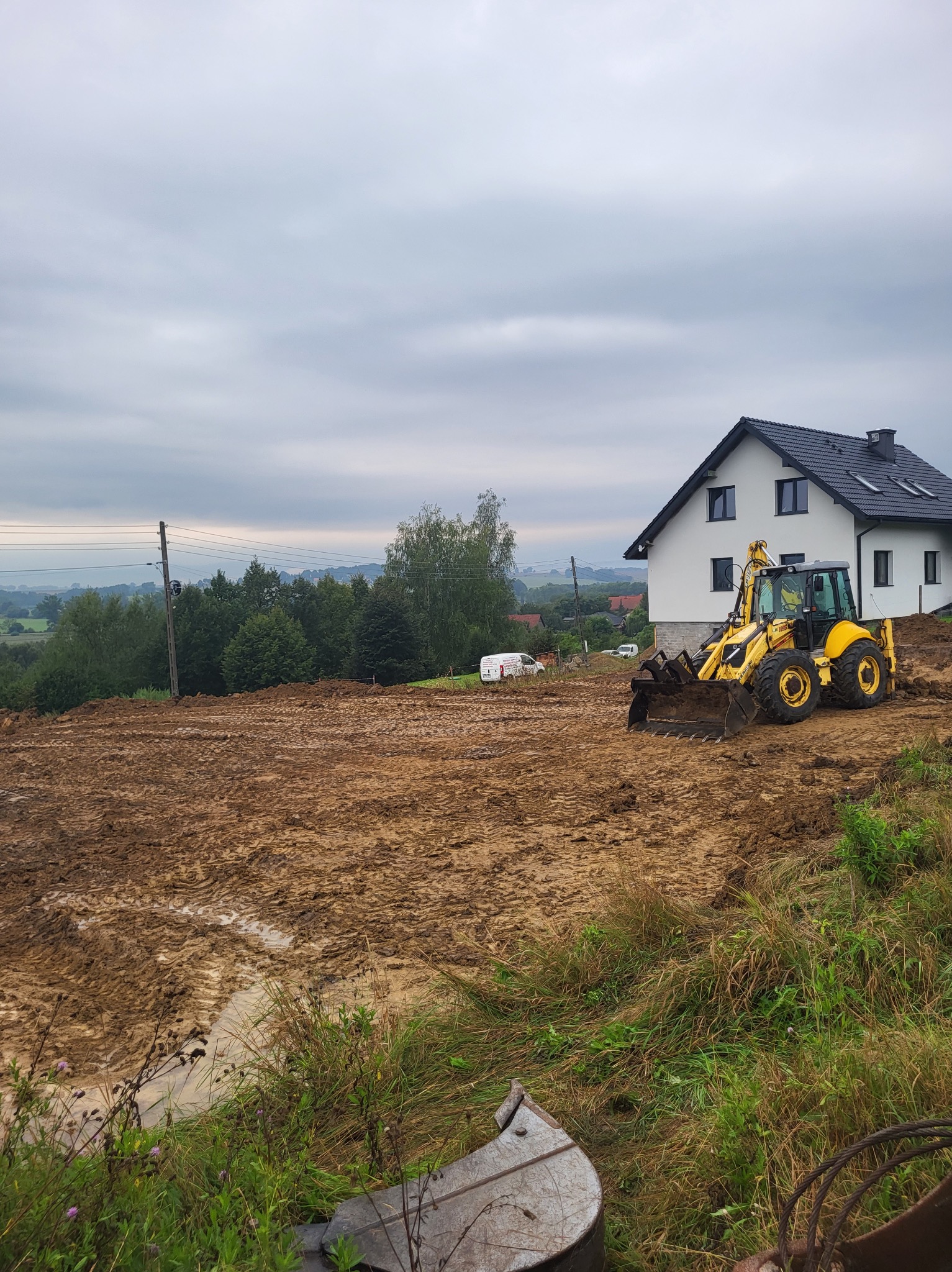 Żółta koparko-ładowarka na placu budowy, przygotowuje teren pod budowę domu jednorodzinnego, widoczne ślady opon w błotnistym gruncie, w tle biały dom z ciemnym dachem i zalesione wzgórza...
