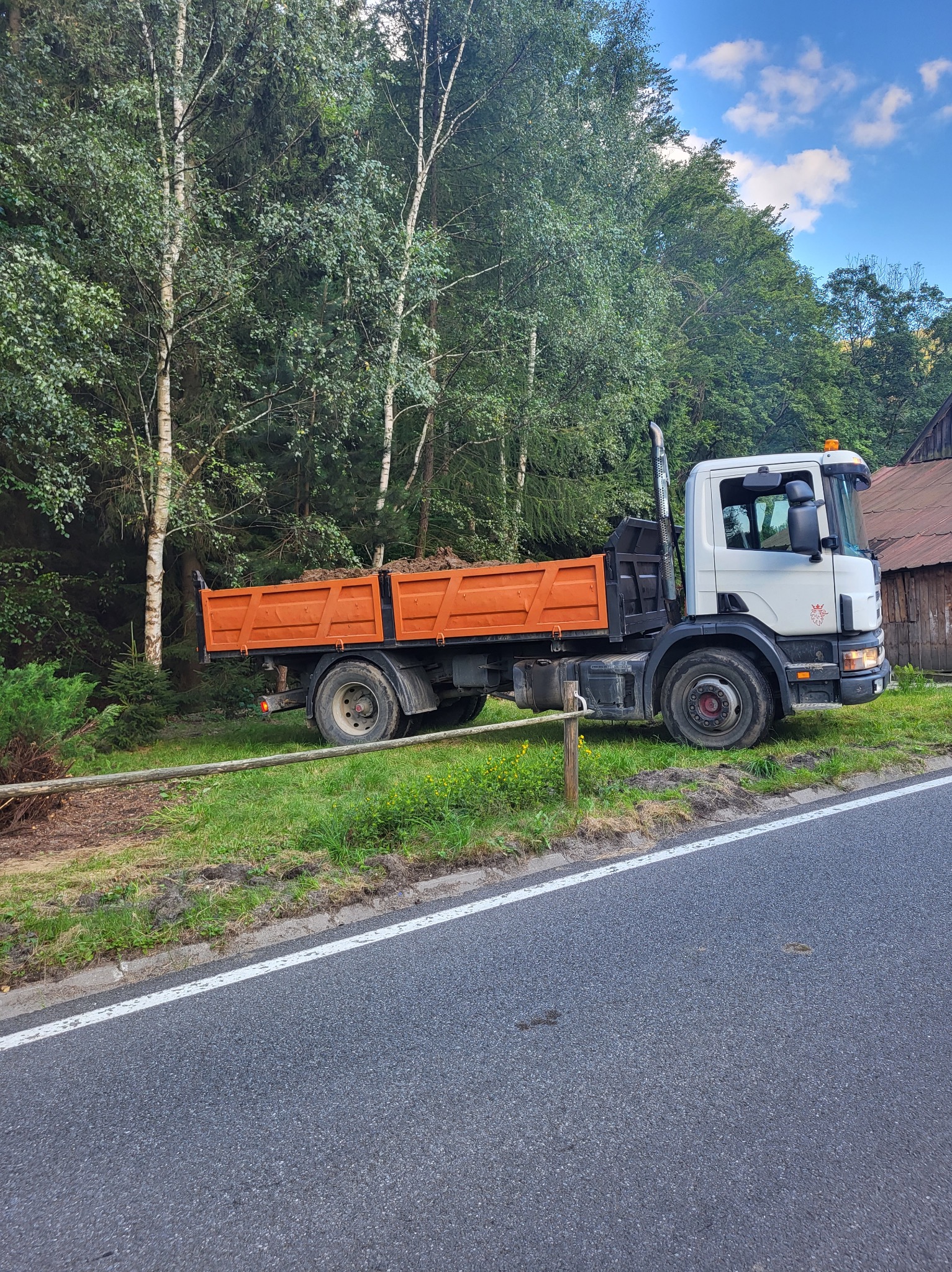 Pomarańczowa wywrotka Scania zaparkowana na poboczu, częściowo za drewnianym płotkiem, z ładunkiem ziemi w skrzyni, na tle zielonego lasu i drewnianego budynku.