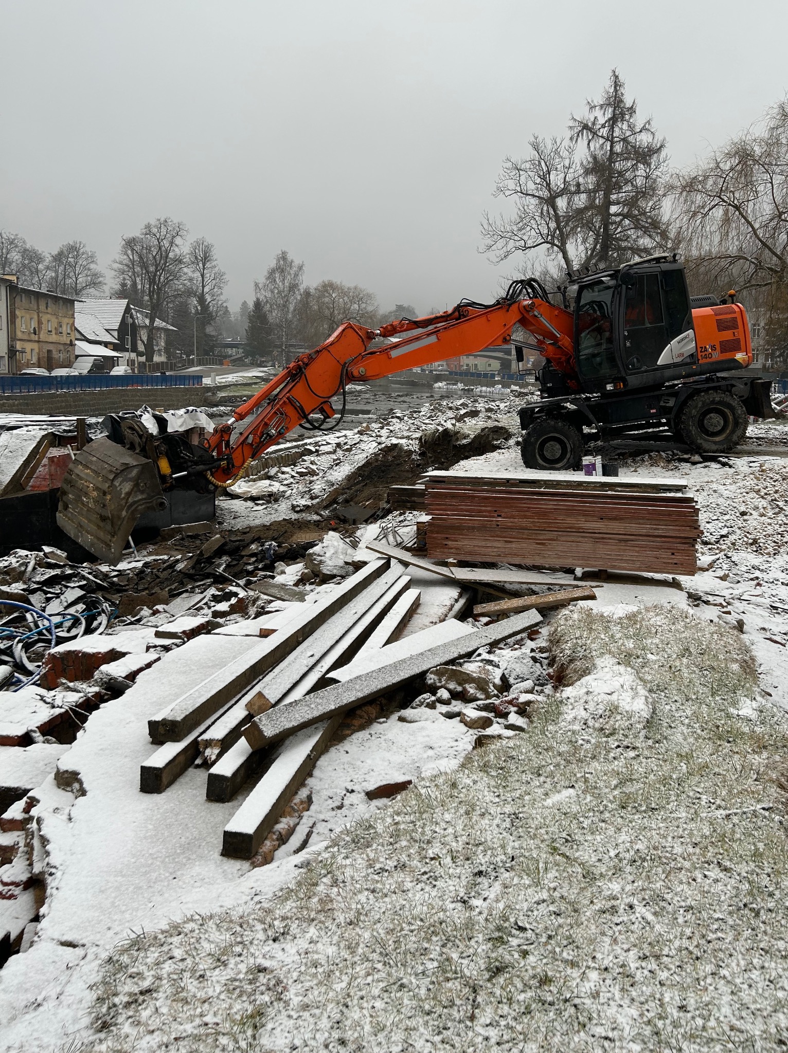 Pomarańczowa koparko-ładowarka na placu budowy w zimowej scenerii, z fragmentami gruzu, drewnianych belek i stertą płyt, częściowo przykrytych śniegiem.
