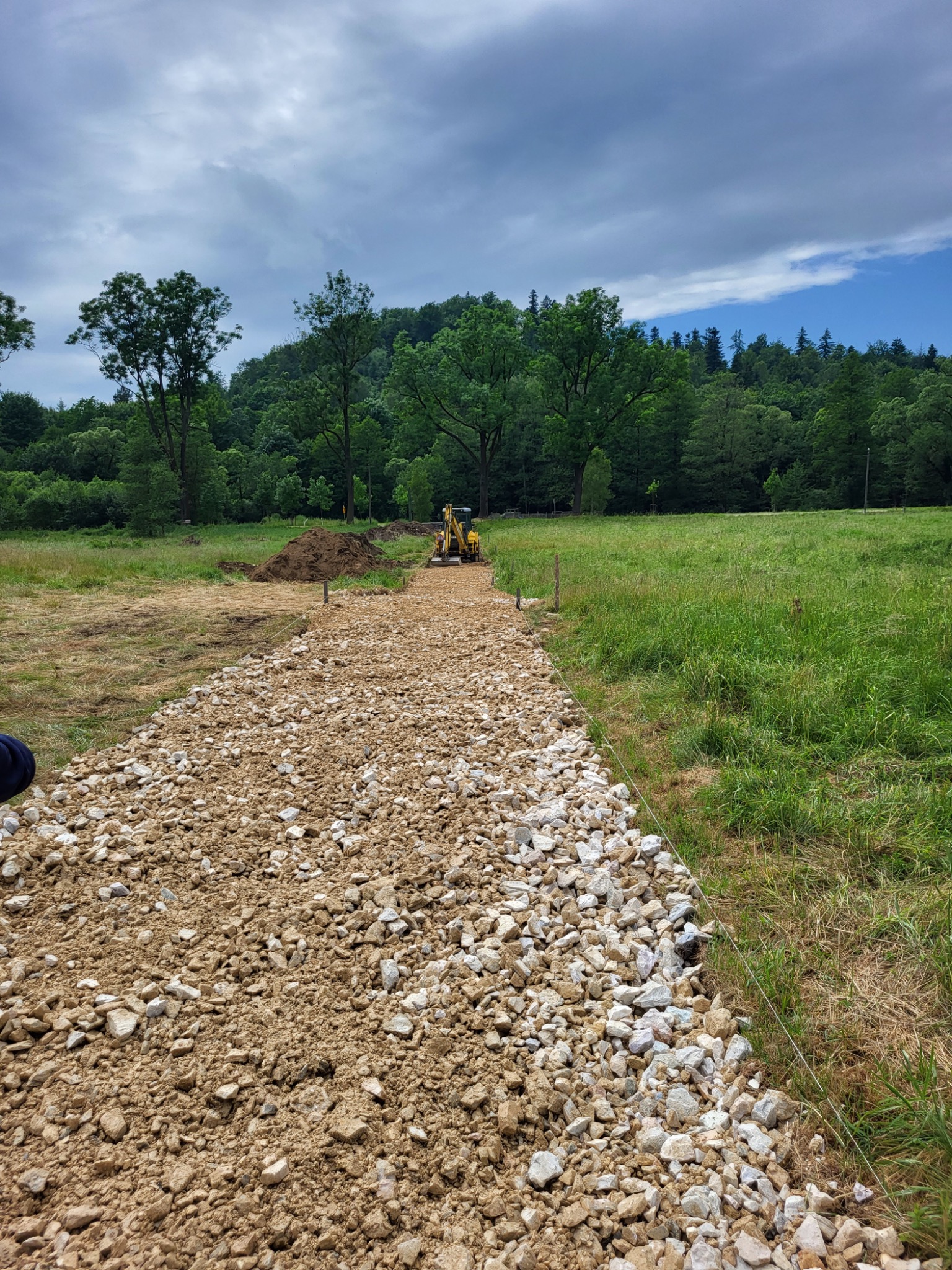 Żwirowa droga w trakcie budowy na łące, w tle las i pracująca koparko-ładowarka, wyznaczona linia granicy pasa żwirowego.