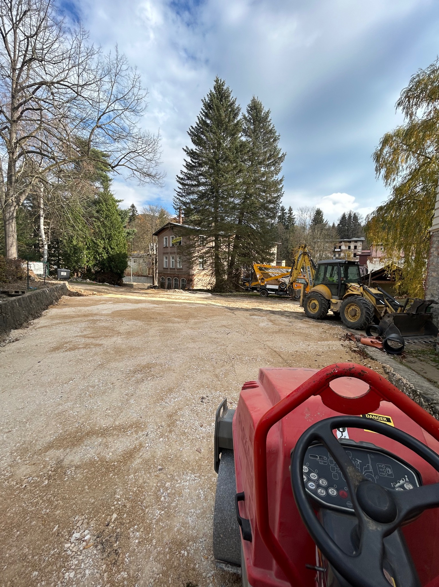 Czerwona zagęszczarka gruntu na pierwszym planie, w tle teren budowy z żółtą koparko-ładowarką i częściowo rozebranym budynkiem w otoczeniu drzew.