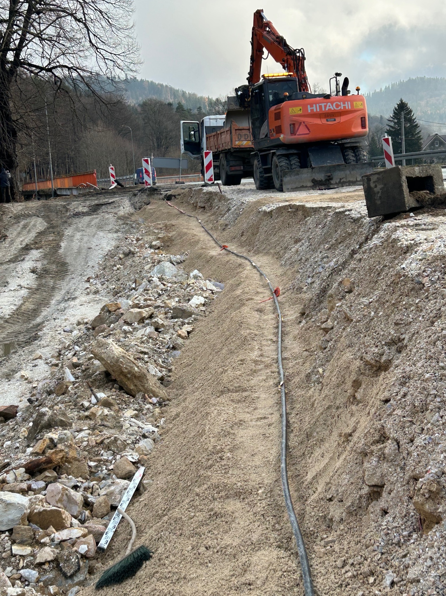 Pomarańczowa koparko-ładowarka Hitachi na tle wykopu pod drogę, z widocznymi elementami infrastruktury i gruzem. W tle góry i drzewa.