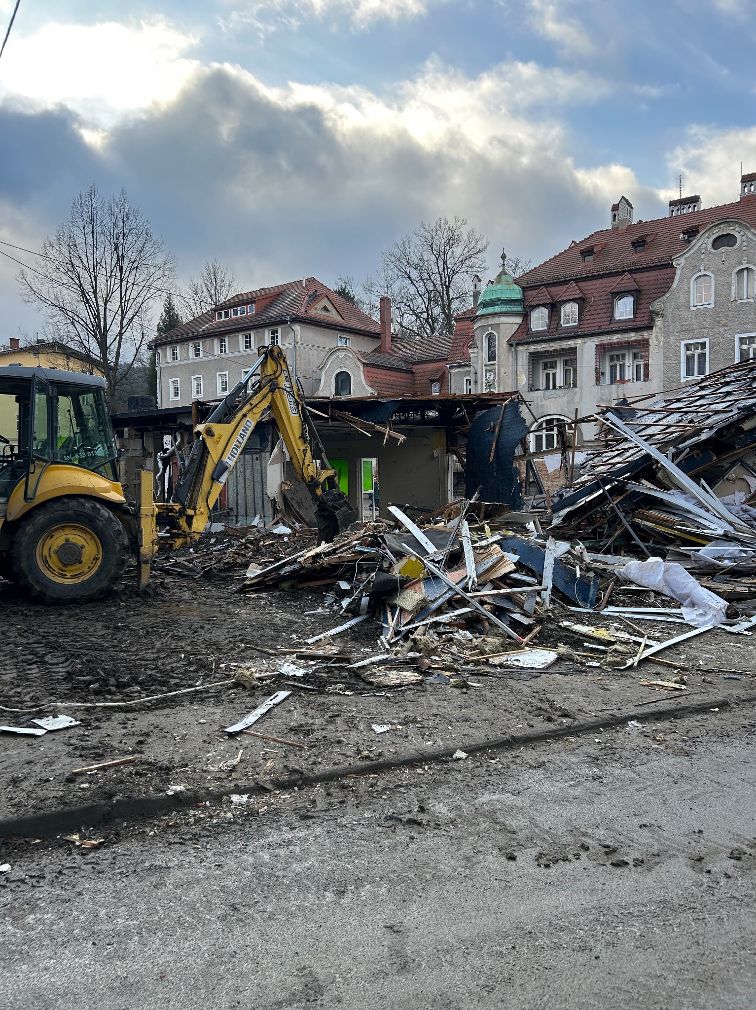 Żółta koparko-ładowarka Holland na placu budowy wśród gruzów po rozbiórce budynku, na tle zabudowy miejskiej i pochmurnego nieba.