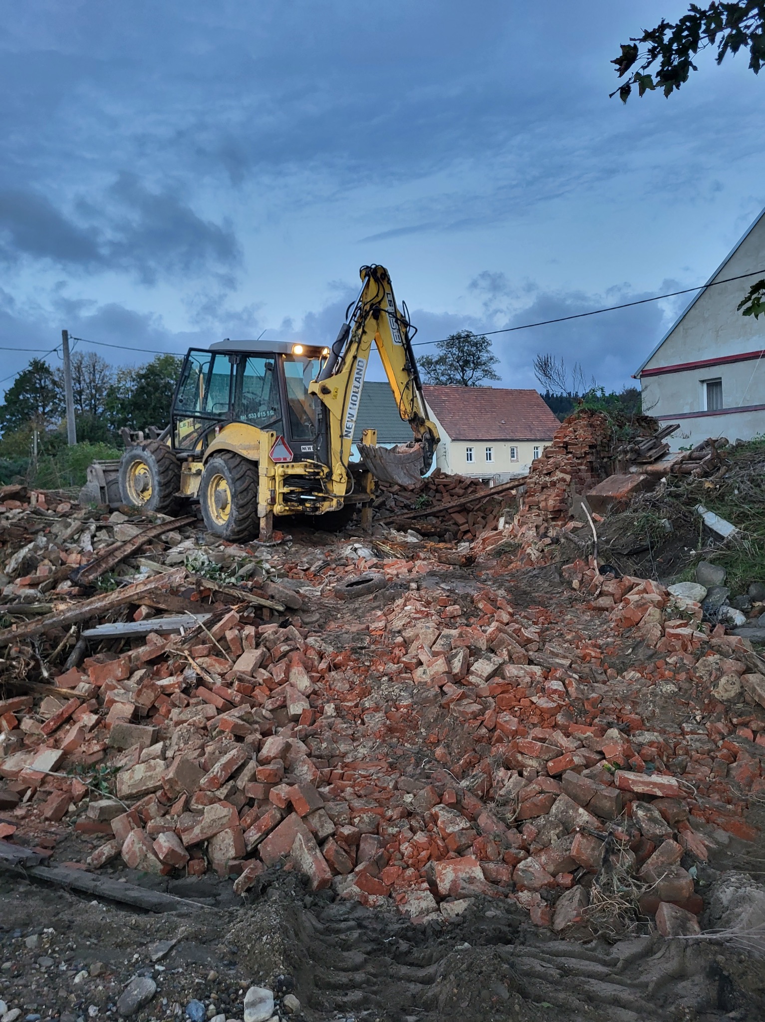 Żółta koparko-ładowarka New Holland na tle sterty gruzu z czerwonej cegły po rozbiórce, z włączonymi światłami, widoczne budynki w tle pod pochmurnym niebem.