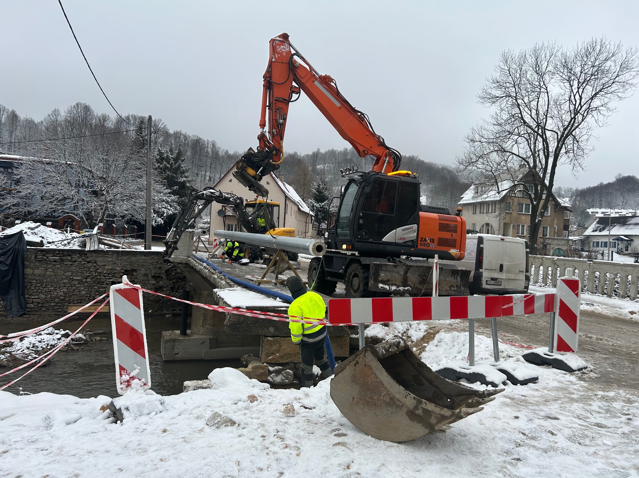 Pomarańczowa koparka Zaxis 140W na zaśnieżonym terenie, prawdopodobnie podczas prac przy rzece lub potoku, z widocznymi barierami ochronnymi, robotnikami w odblaskowych kamizelkach i fragmentami rur.