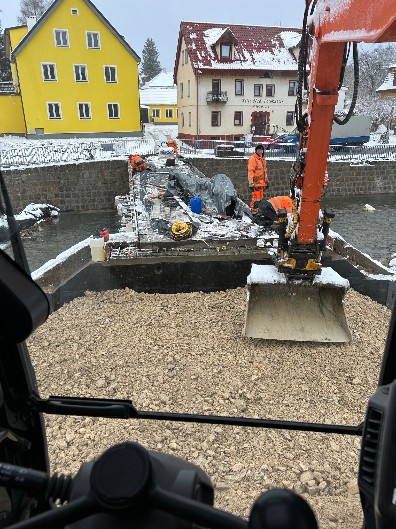 Widok z kabiny koparki na barkę z gruzem na rzece, w tle pracownicy w pomarańczowych kombinezonach i zabudowania w zimowej scenerii.