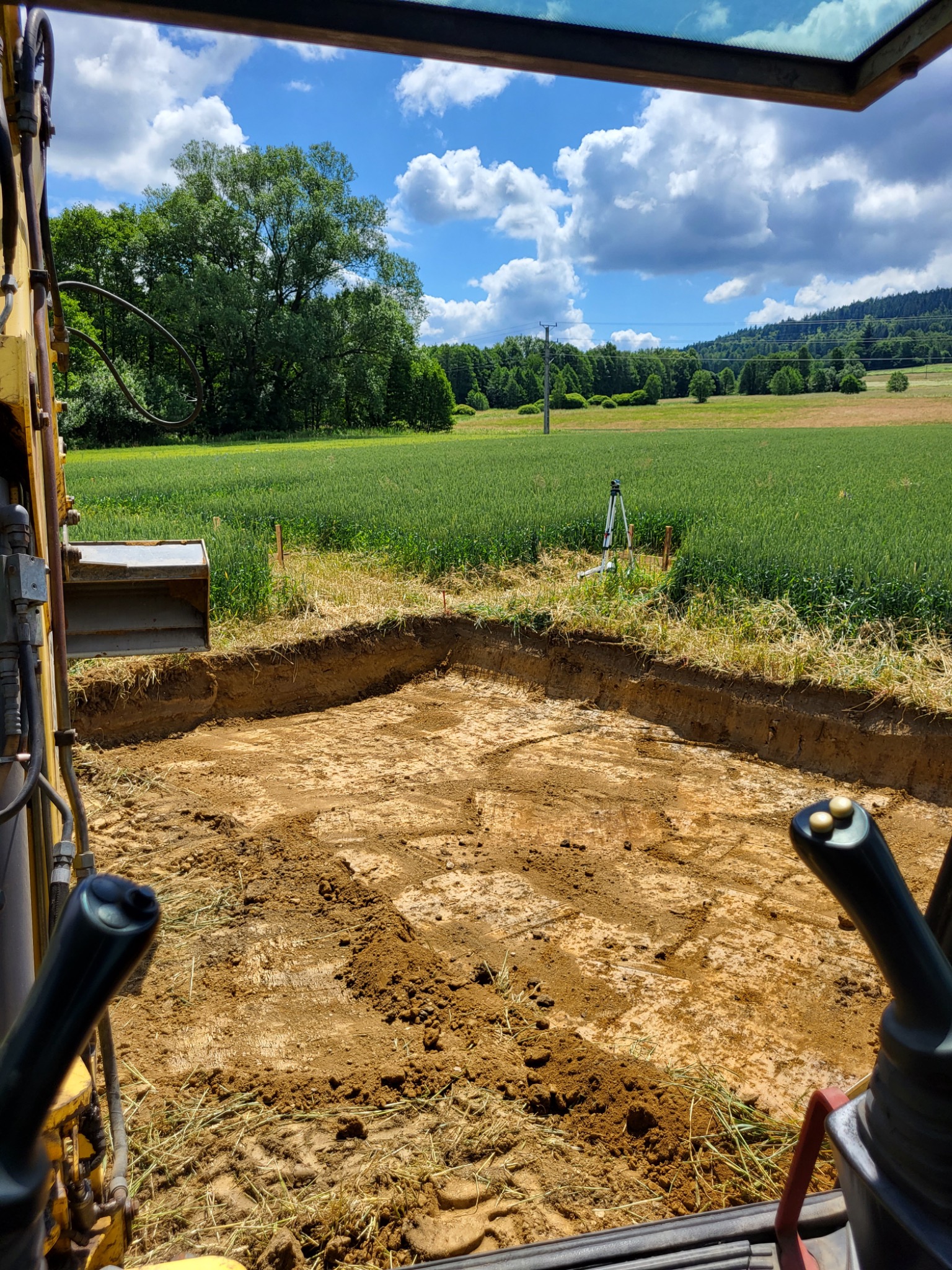 Widok z kabiny koparki na wykop w kształcie litery L, z widocznym niwelatorem laserowym na tle pola i lasu w słoneczny dzień.