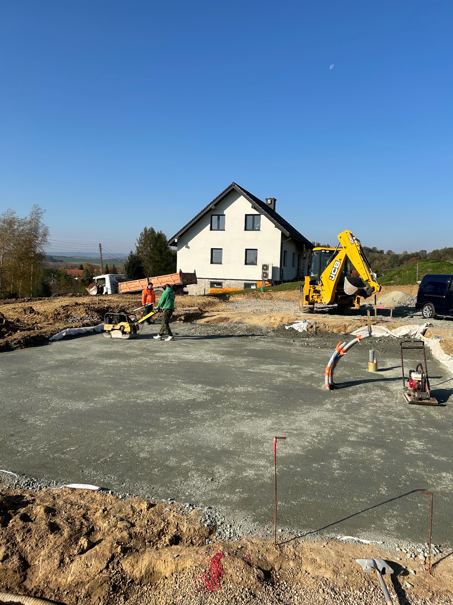 Wylany beton pod fundamenty domu jednorodzinnego, widoczna żółta koparko-ładowarka JCB, ubijarka, pracownicy, wykopane fundamenty, niebieskie niebo i krajobraz w tle.