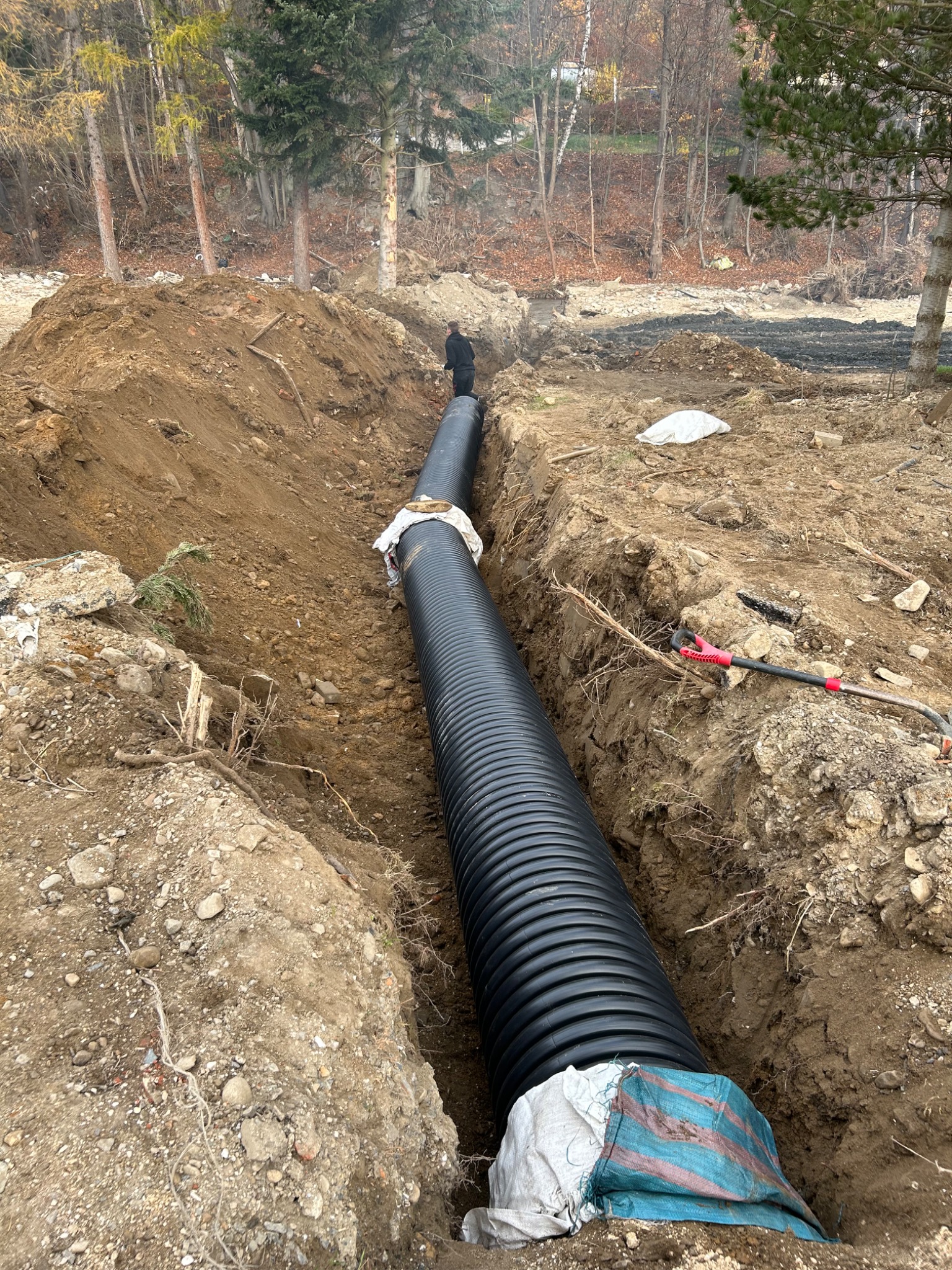 Długi odcinek czarnej, karbowanej rury ułożonej w głębokim wykopie w ziemi, zabezpieczony workami; w tle widoczna postać w ciemnym ubraniu i drzewa.