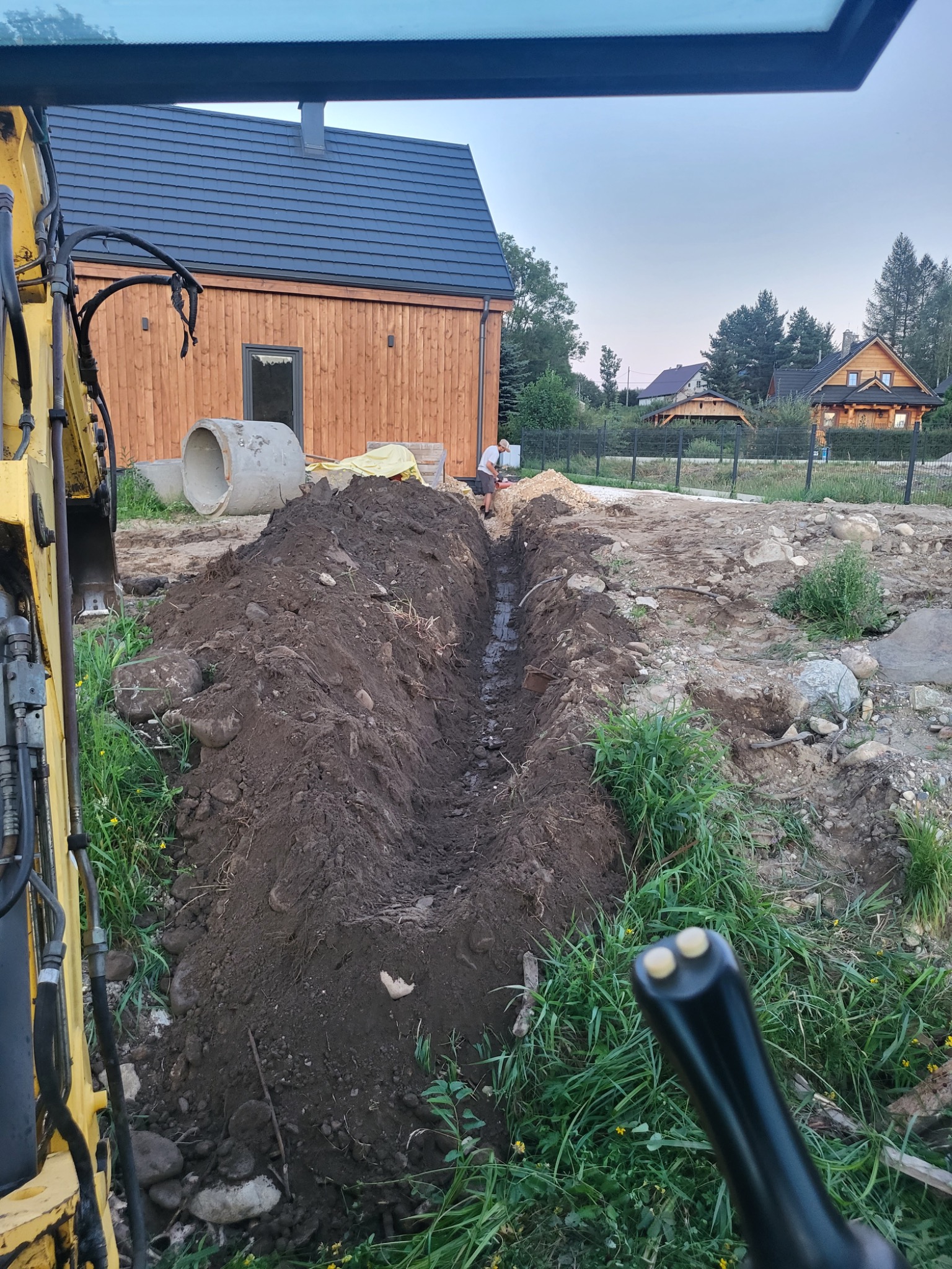 Wykopany wąski rów w ziemi z widoczną koparką z boku, w tle budynek mieszkalny z drewnianą elewacją i betonowe rury oraz pracownik. Ujęcie z kabiny koparki.