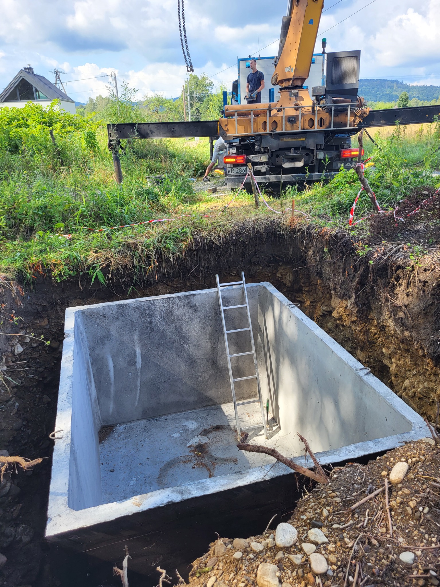Betonowy zbiornik z drabinką w wykopie, opuszczany na łańcuchach dźwigu, w tle pracownik obsługujący dźwig, teren budowy zabezpieczony taśmą.