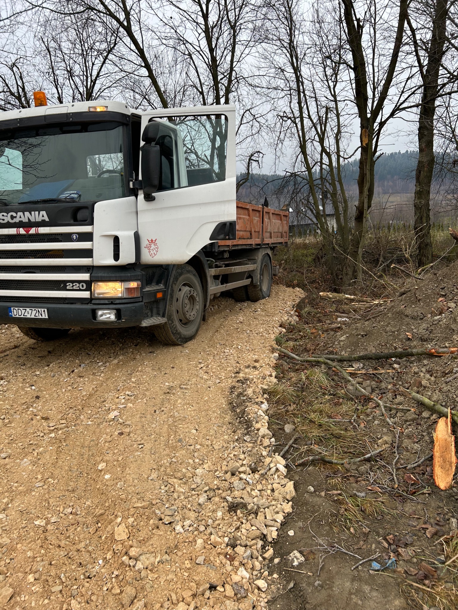 Ciężarówka Scania 220 na żwirowej drodze gruntowej, z otwartymi drzwiami kierowcy, widoczny fragment wykarczowanego terenu i drzewa w tle.