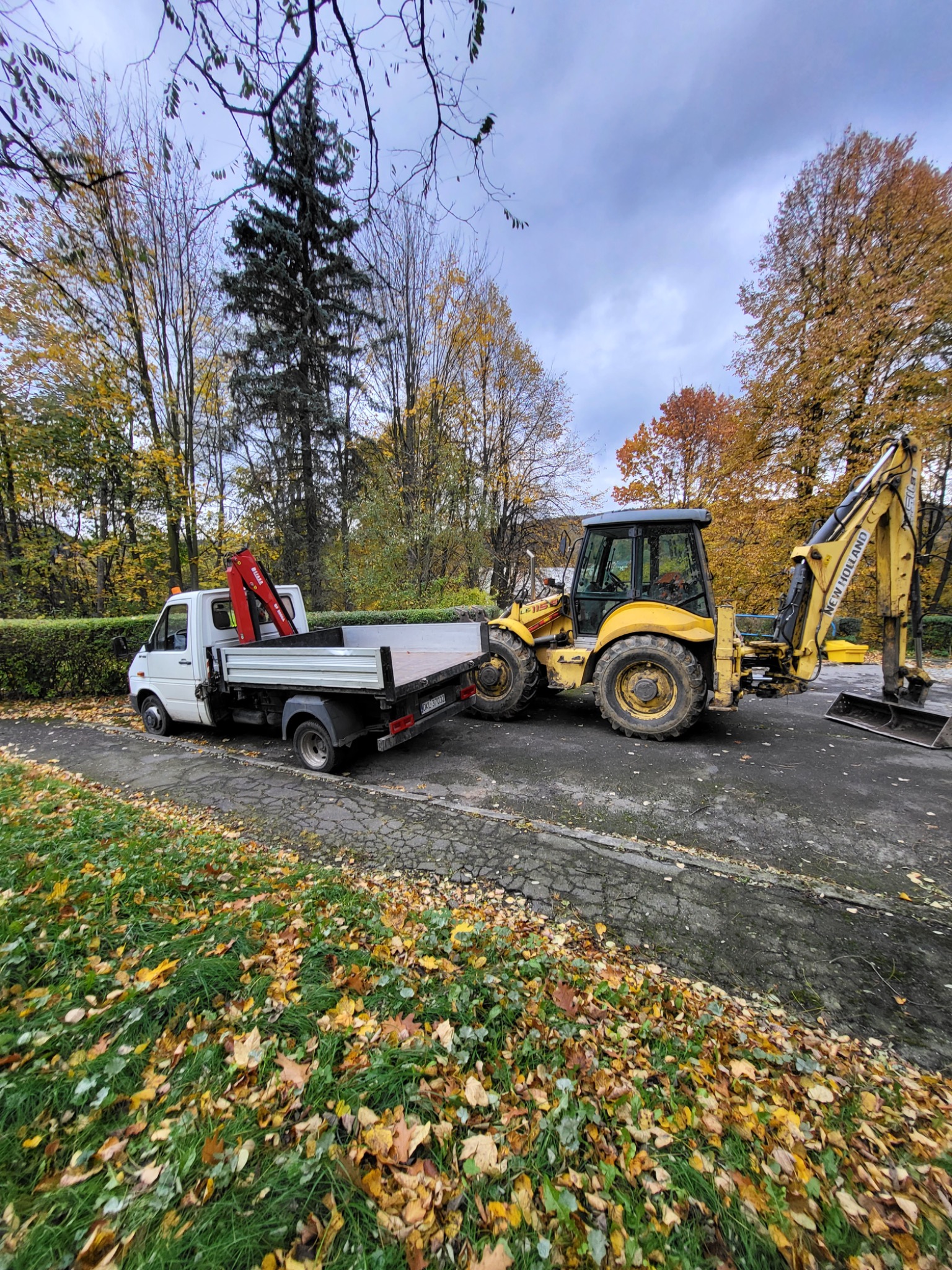 Żółta koparko-ładowarka New Holland i biały samochód dostawczy z dźwigiem na tle jesiennego krajobrazu z drzewami w parku.
