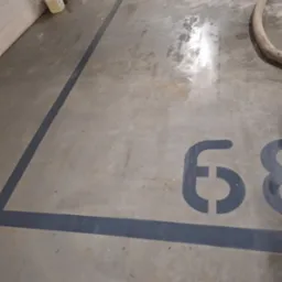 Wilgotna posadzka betonowa z namalowanym numerem '68' wewnątrz czarnych linii wyznaczających stanowisko postojowe, widoczny wąż i kanister z płynem.