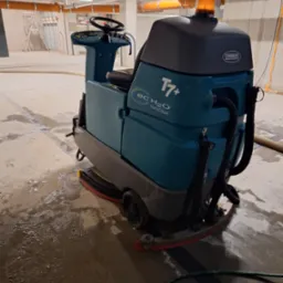 Automat szorująco-zbierający Tennant T7+ NanoClean na mokrej, betonowej posadzce w hali w trakcie sprzątania, widoczne ślady po czyszczeniu, pomarańczowe rury pod sufitem.