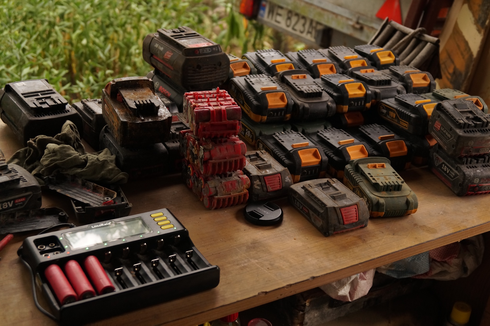 Stół warsztatowy zastawiony bateriami do elektronarzędzi różnych marek, w tym Bosch i Teccpo, oraz ładowarką LitoKala z trzema czerwonymi akumulatorami w trakcie ładowania. Niektóre baterie są...