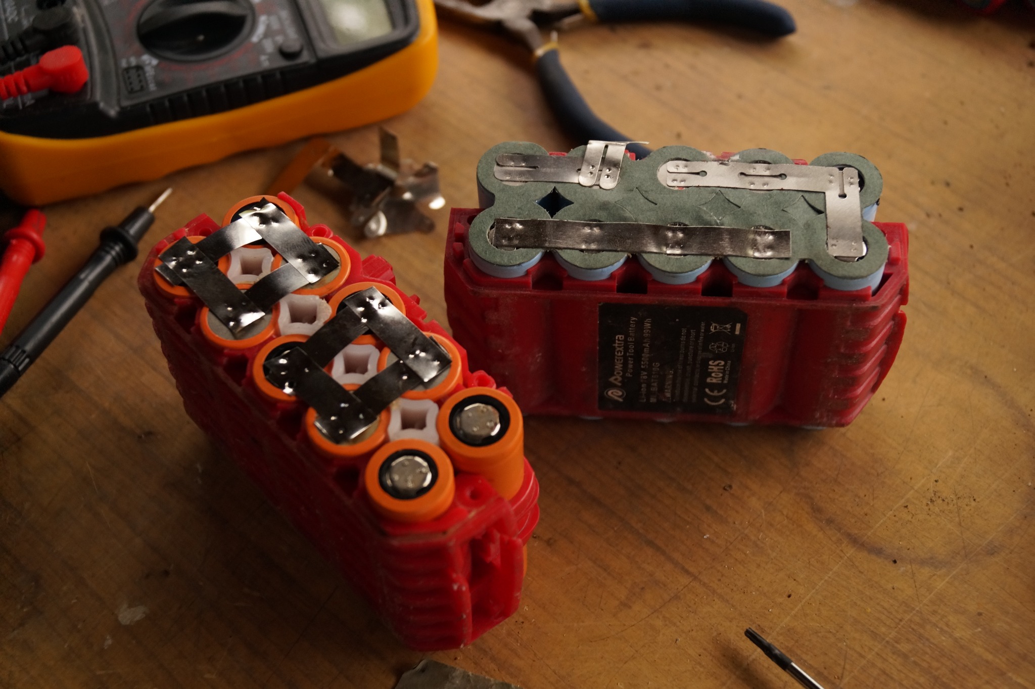 Dwa czerwone, częściowo zdemontowane akumulatory elektronarzędzi na drewnianym stole, widoczne elementy mocujące i ogniwa, w tle multimetr i szczypce.