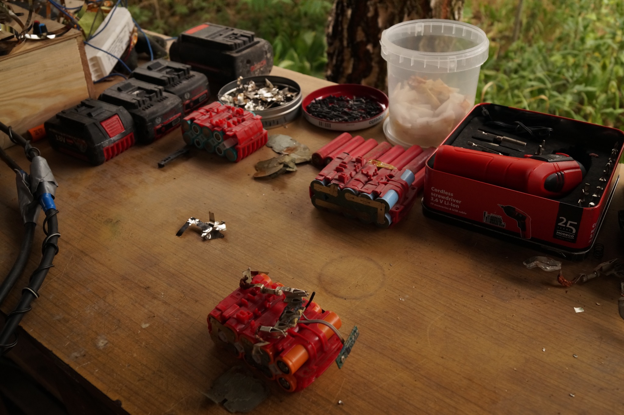 Warsztatowy stół z rozłożonymi akumulatorami do elektronarzędzi w trakcie naprawy, widoczne narzędzia, lutownica, oraz pojemniki z elementami do naprawy.